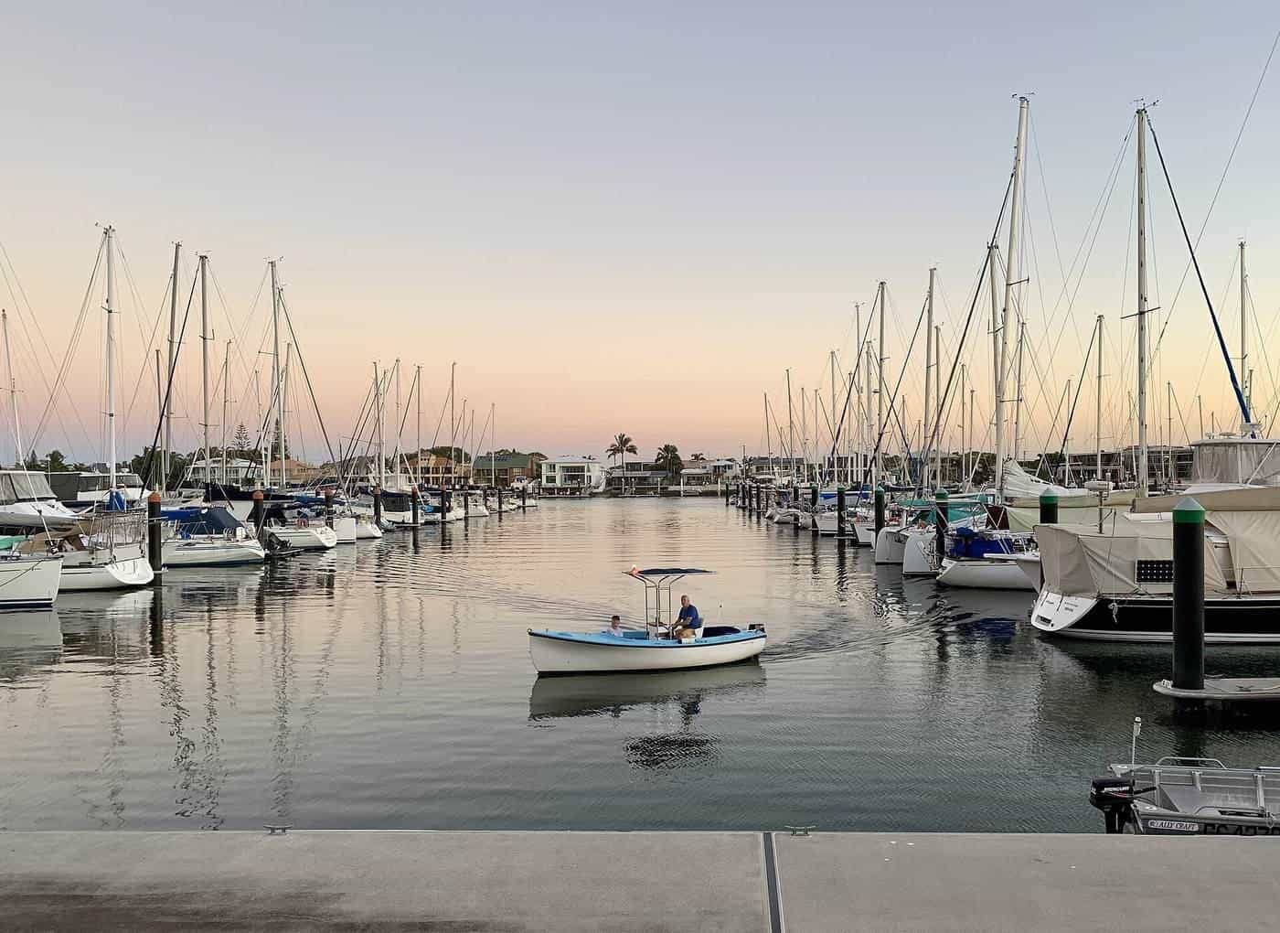 Rows of yachts tied up at a marina at sunrise, with a small motorboat crossing the water
